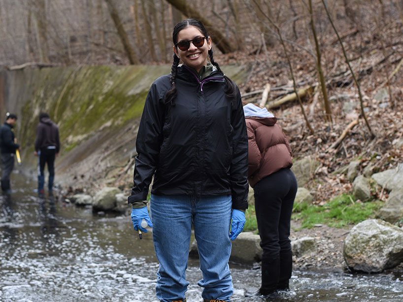 Kelsey Hazel smiling, standing in a forest in a shallow river, with knee high rubber boots.