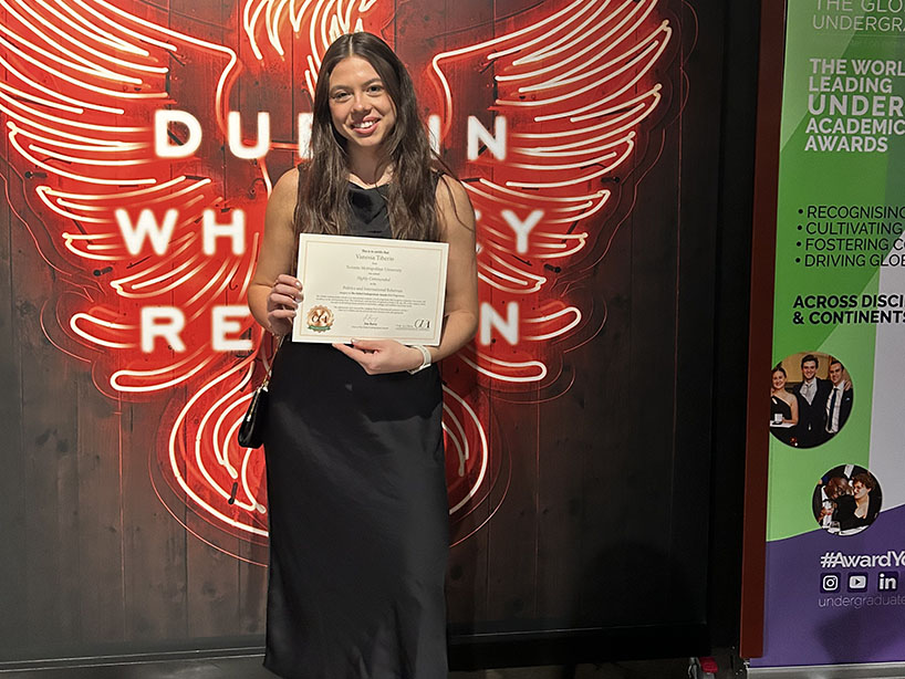  Vanessa Tiberio holding her award certificate in front of a neon sign at the Global Undergraduate Award ceremony.