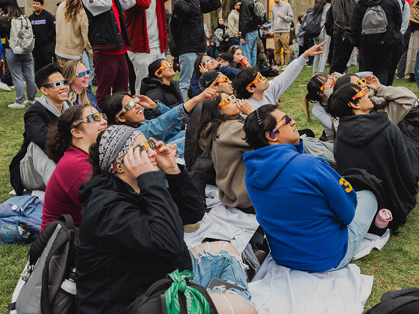 A group of attendees at TMU’s Solar Eclipse Watch Party, wearing protective glasses, point up and look at the sky.