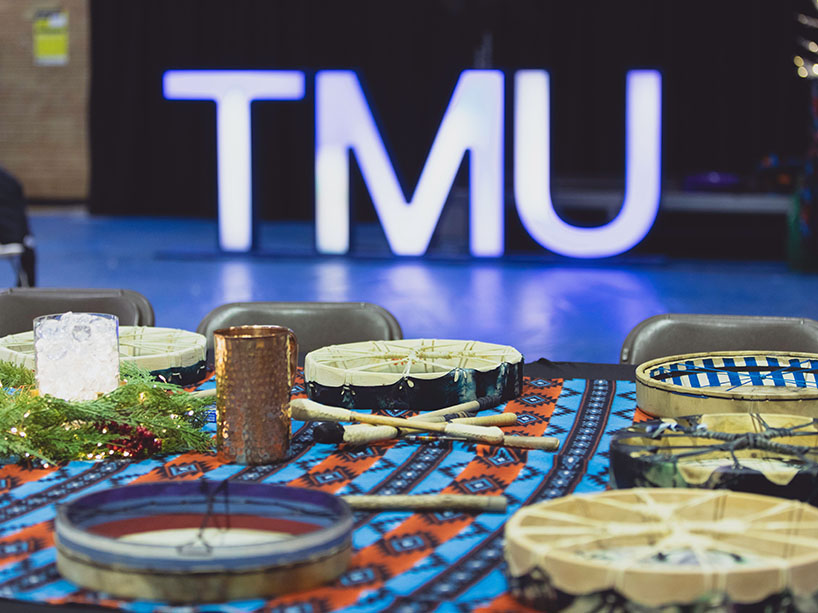 A table of Indigenous drums sits in front of a lit up TMU sign