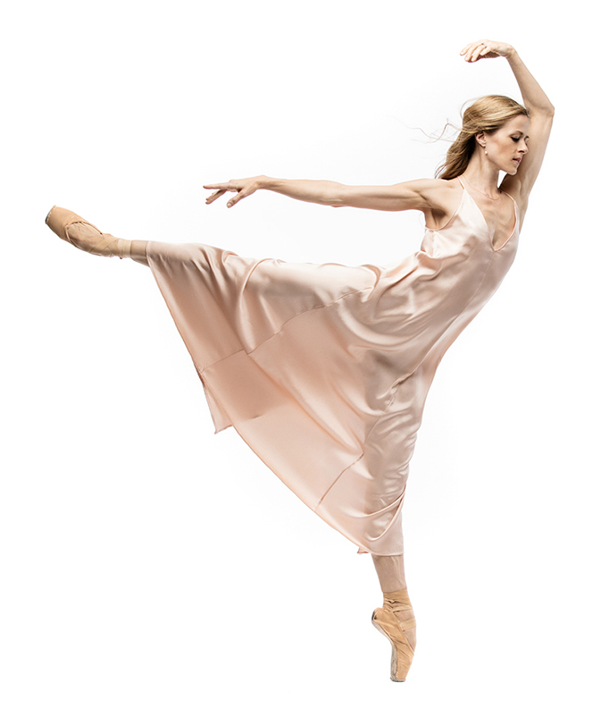 In a pale pink satin dress, Heather Ogden stands on pointe with 1 leg extended behind her.