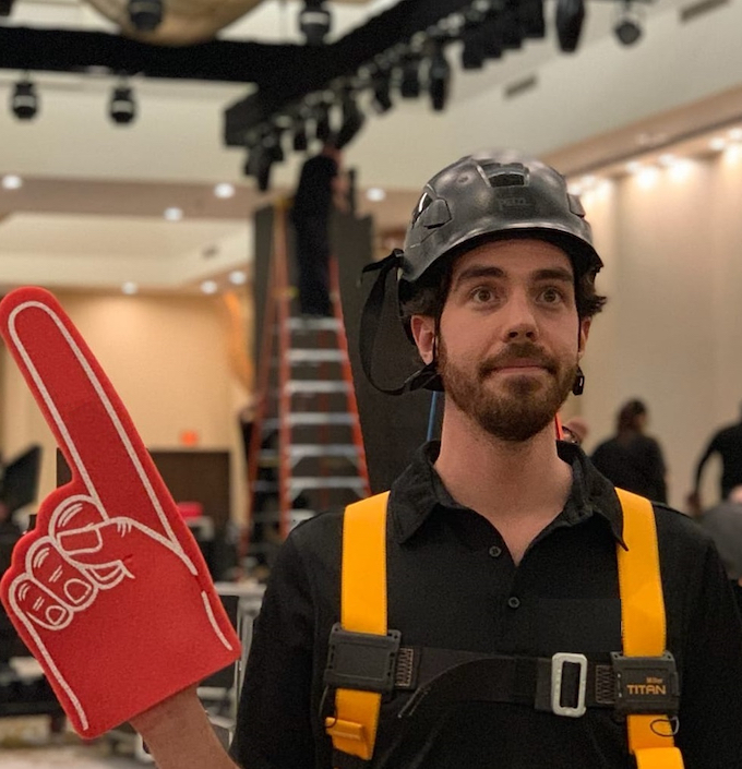 A person in a hard hat and harness wears a foam finger