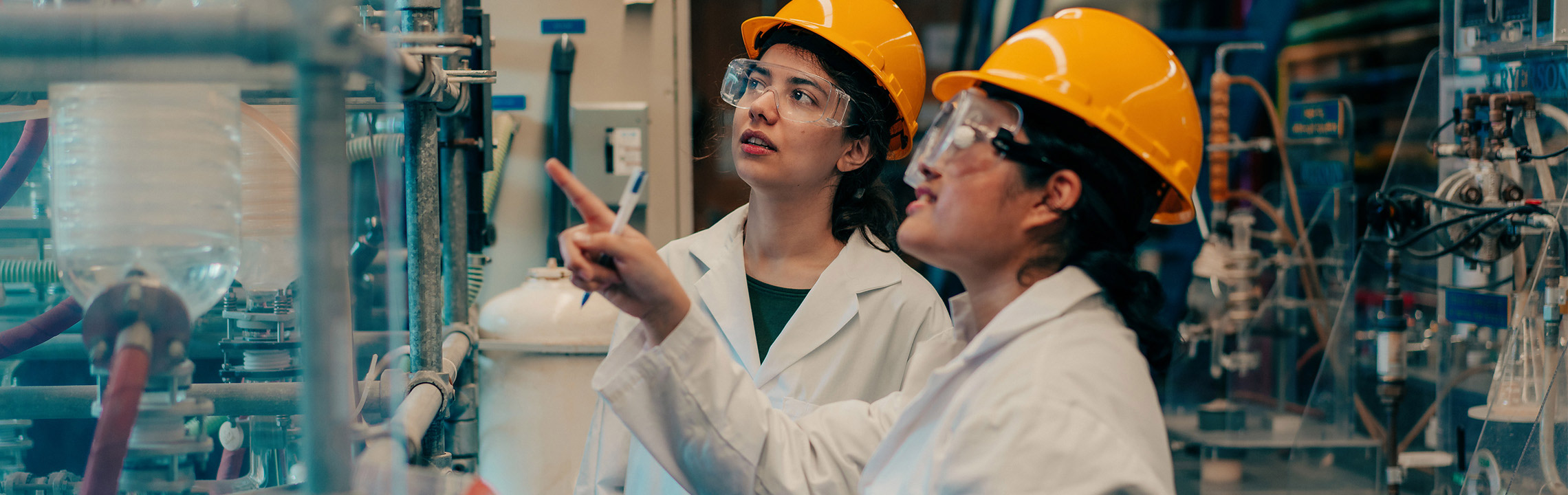 Two Chemical Engineering students in hard hats, googles and lab coats discuss a project together.