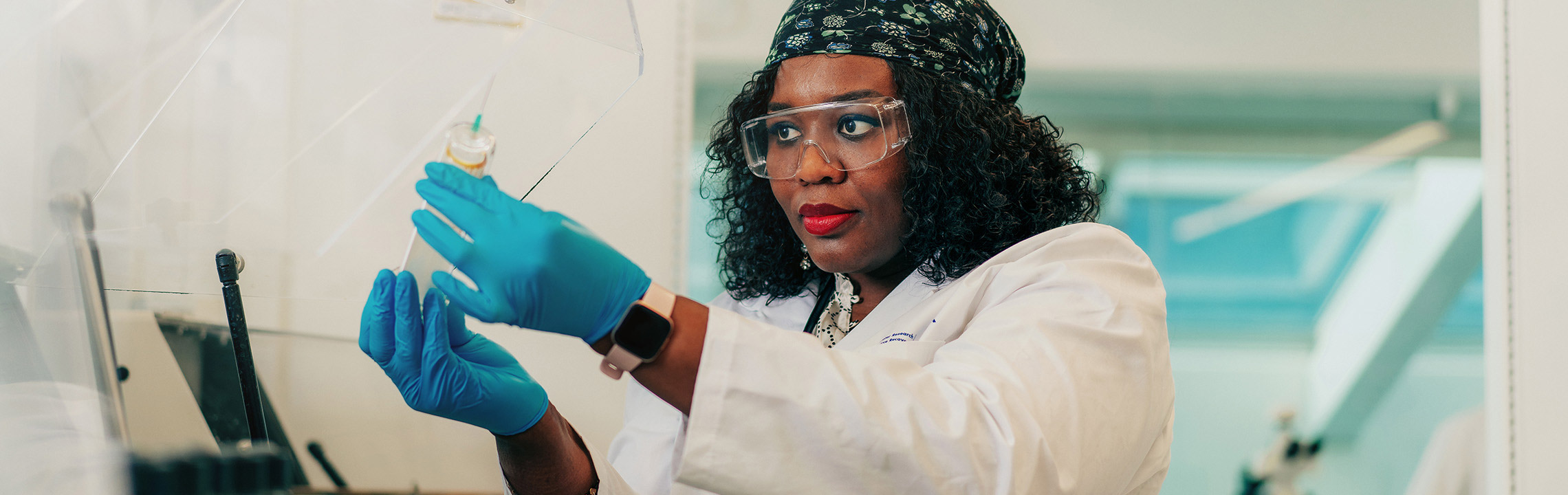 A Civil Engineering students in a lab coat, googles and gloves inspects a project in a lab.