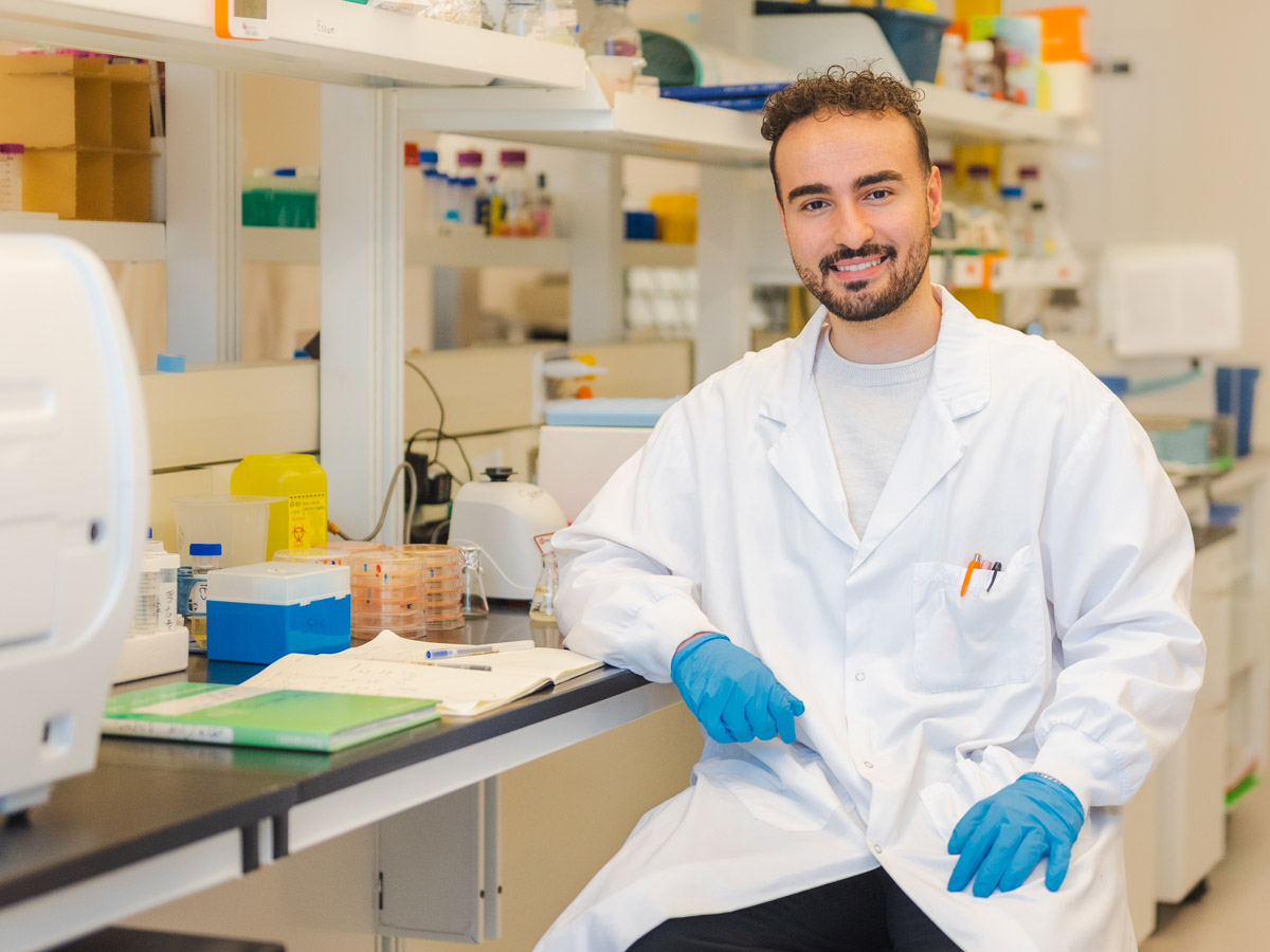 Essam Karam sitting at a lab bench wearing a lab coat and blue latex gloves.