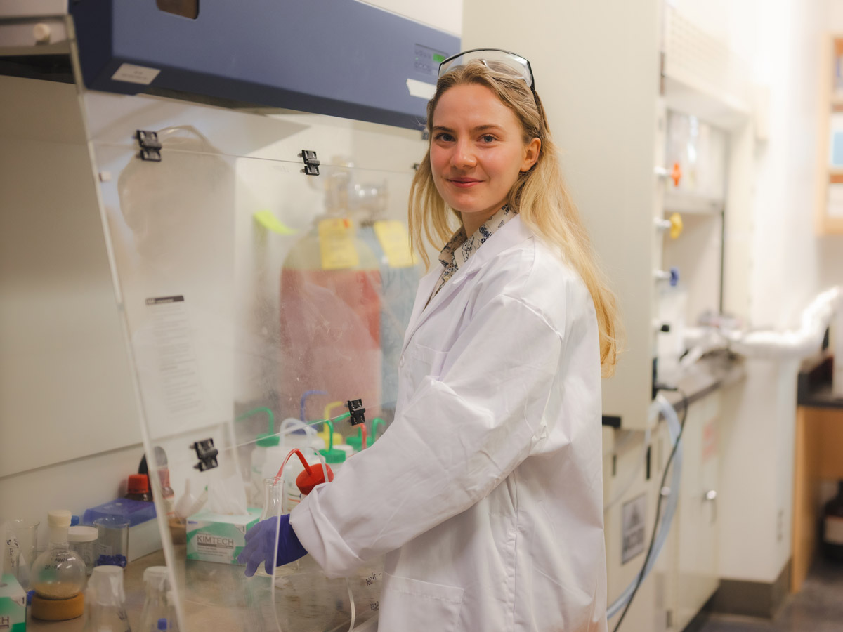 Reese Grandy in a lab coat, goggles and gloves working in a lab.