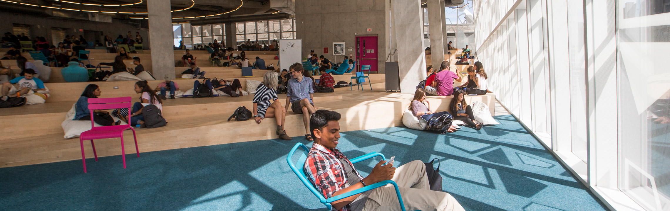 A student smiles at his phone while lounging by the sunny windows of the Student Learning Centre (SLC) "Beach" floor.