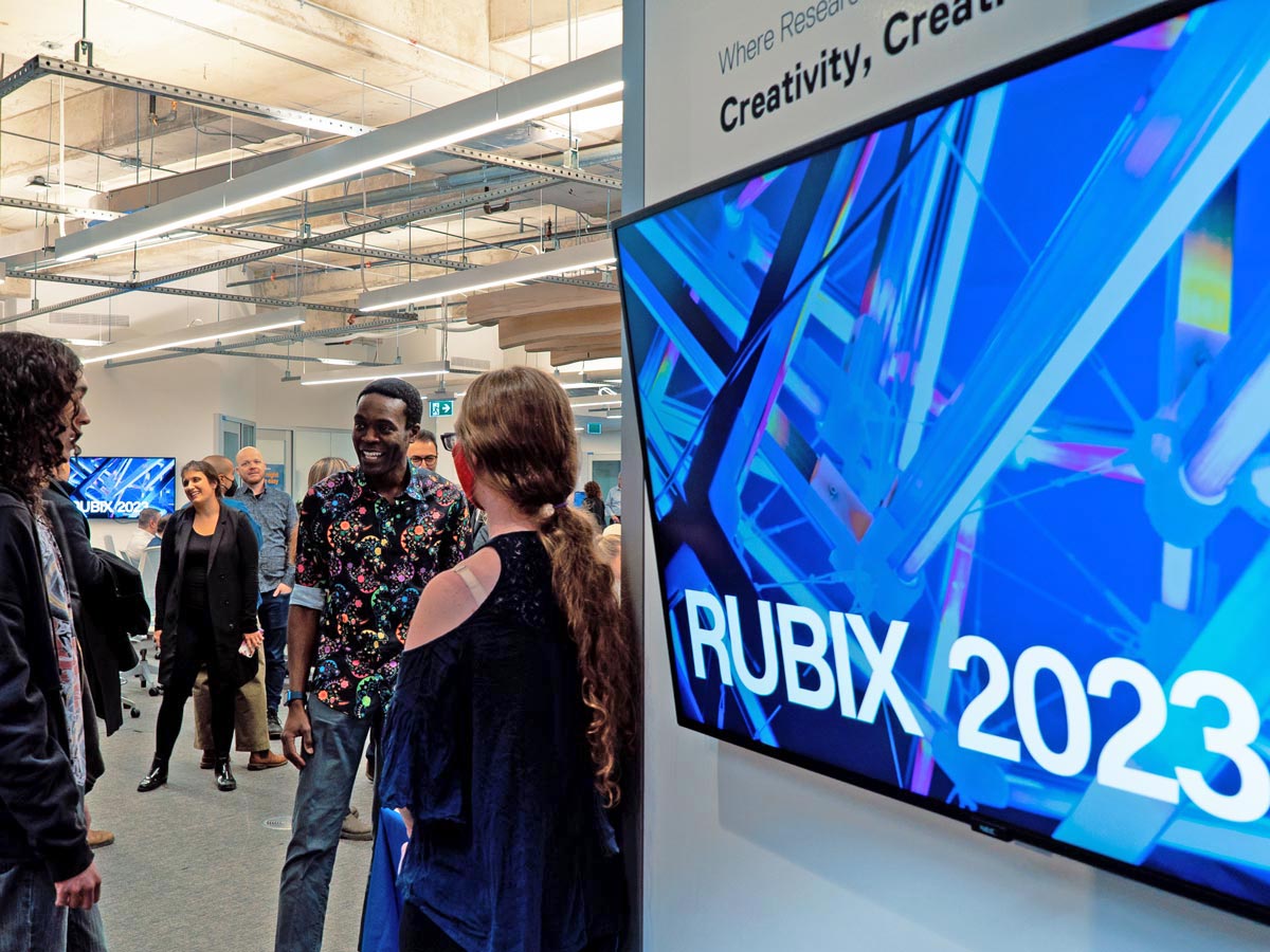 A screen with an image showing connected wires and beams in blue welcomes attendees to RUBIX 2023. Beyond, groups of people can be seen chatting. 