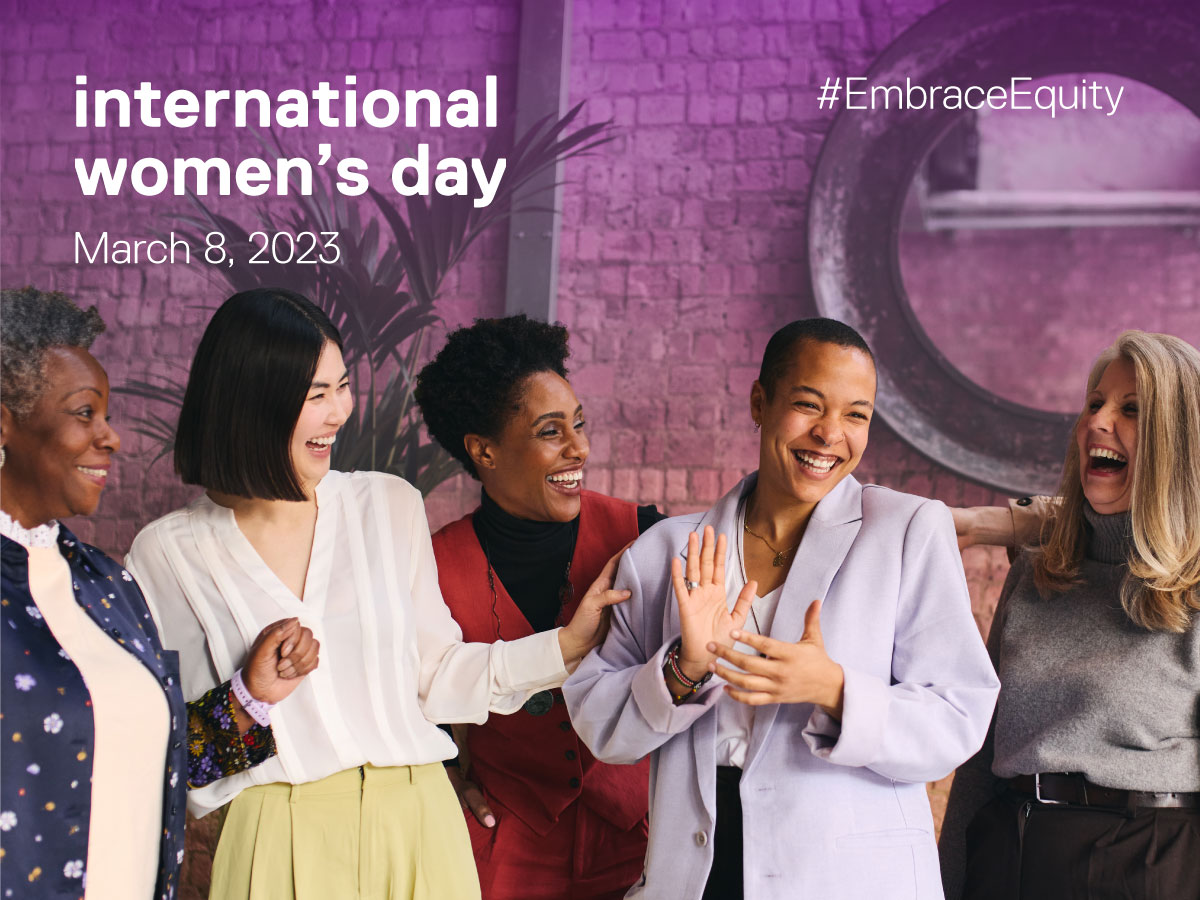 A group of six women laugh together while standing in front of a purple stained background. In white text above their heads it says international women’s day March 8, 2023 and on the right of that it says #EmbraceEquity.