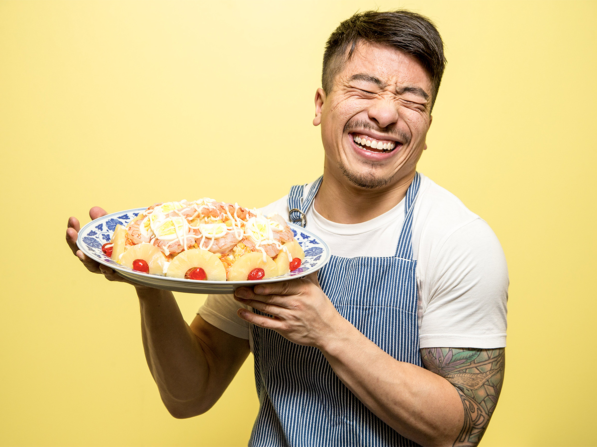 Karon Liu in a white t-shirt, blue striped apron smiling while holding a plate of food which includes pineapples, red cherries and eggs. 