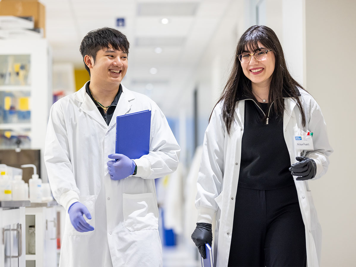 Two science students in lab coats smiling and walking in a lab