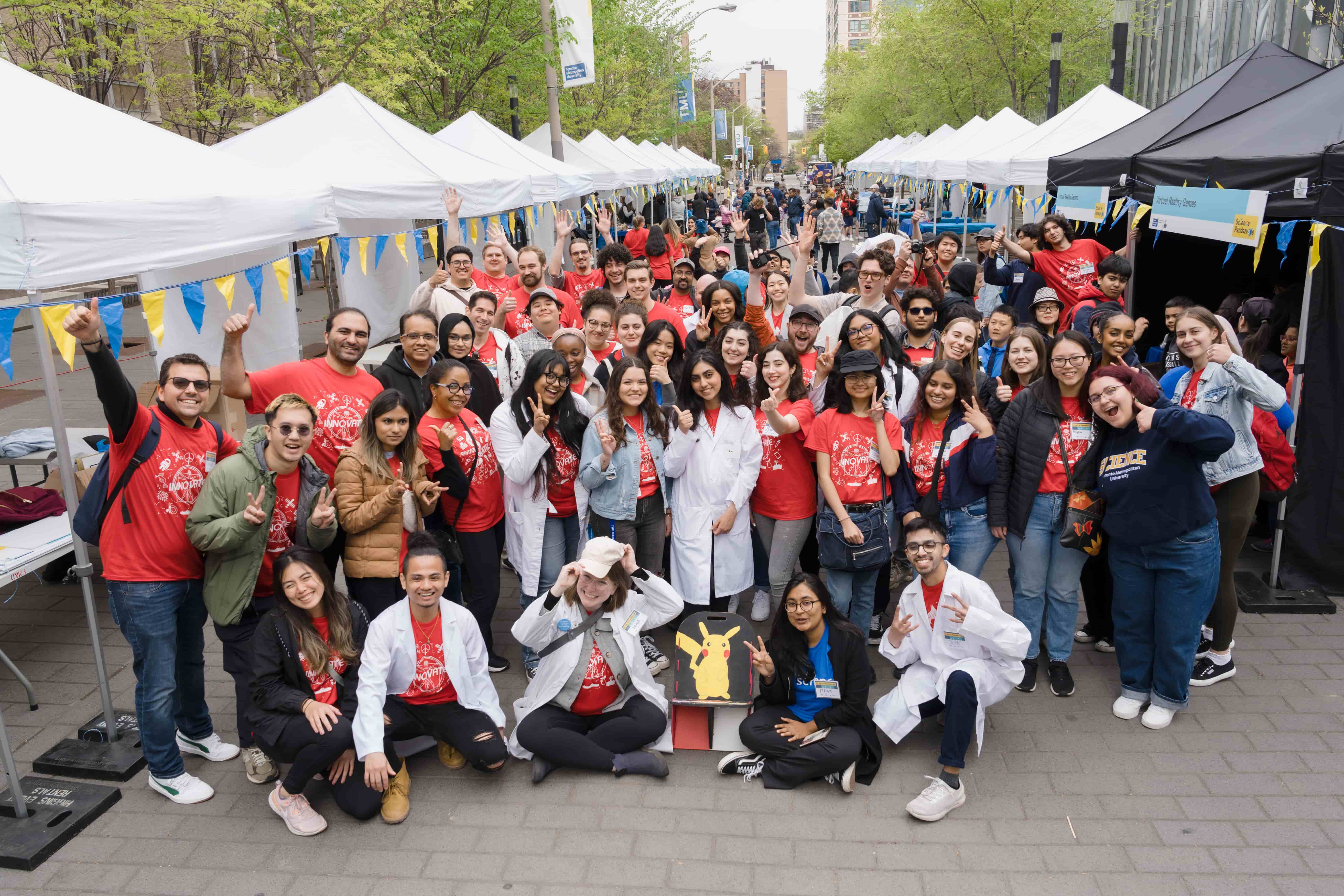Group of about 50 people, wearing red t-shirts, posing for fun, with white event tents in the background