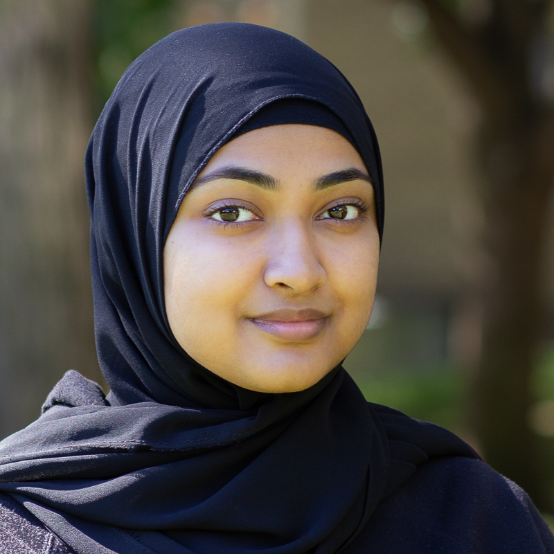 Headshot of Yusra Saifuddin, wearing a navy headscarf, smiling gently