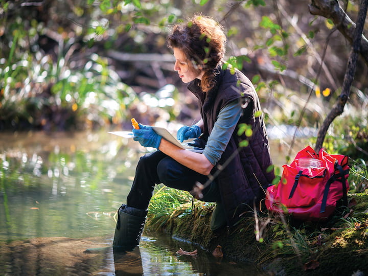 A researcher tests water by a stream, wearing gloves and boots, with a red backpack by her side.