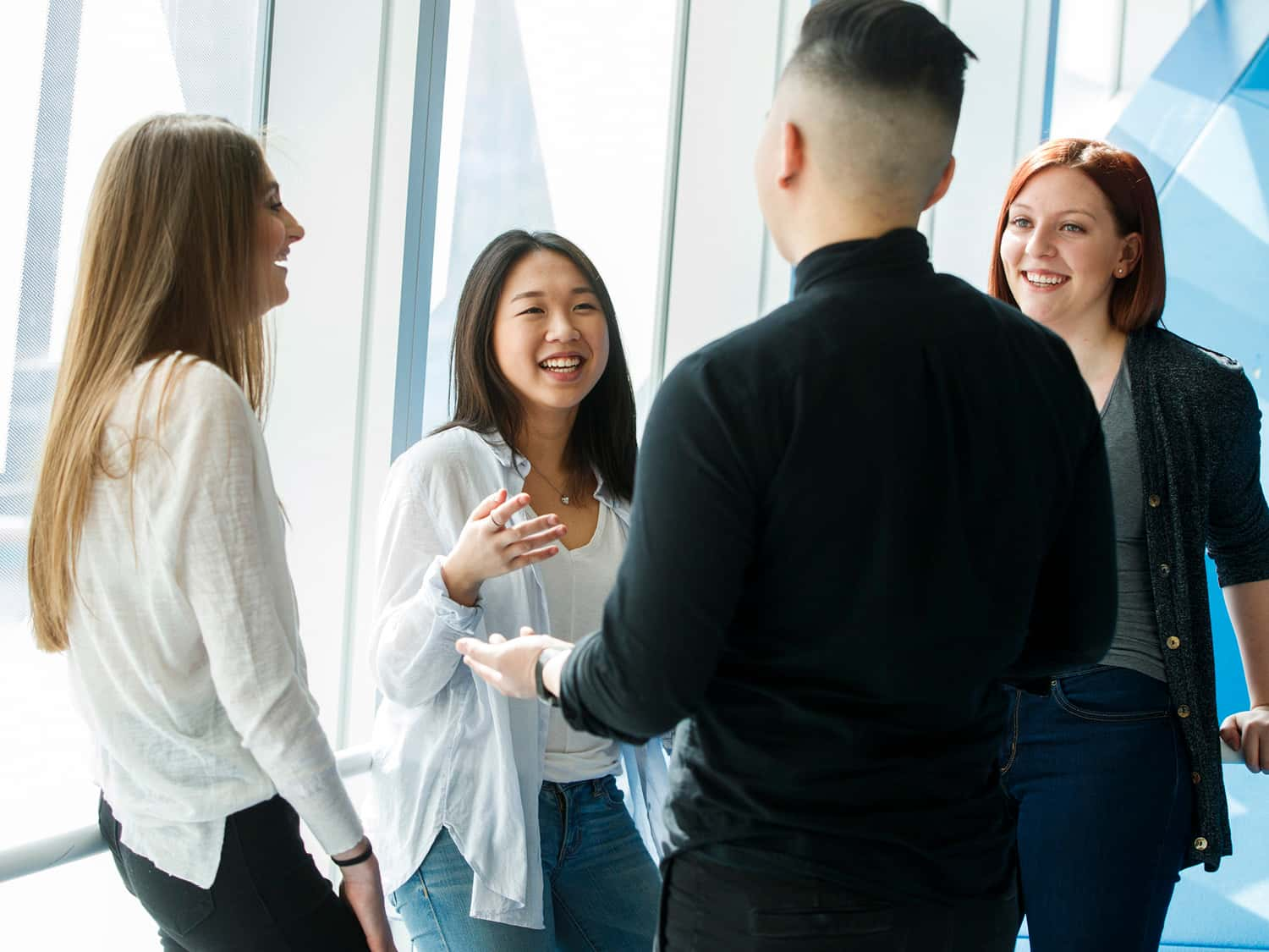 A group of people engaging in a friendly conversation near a large window, with natural light streaming in. They appear relaxed and are smiling.