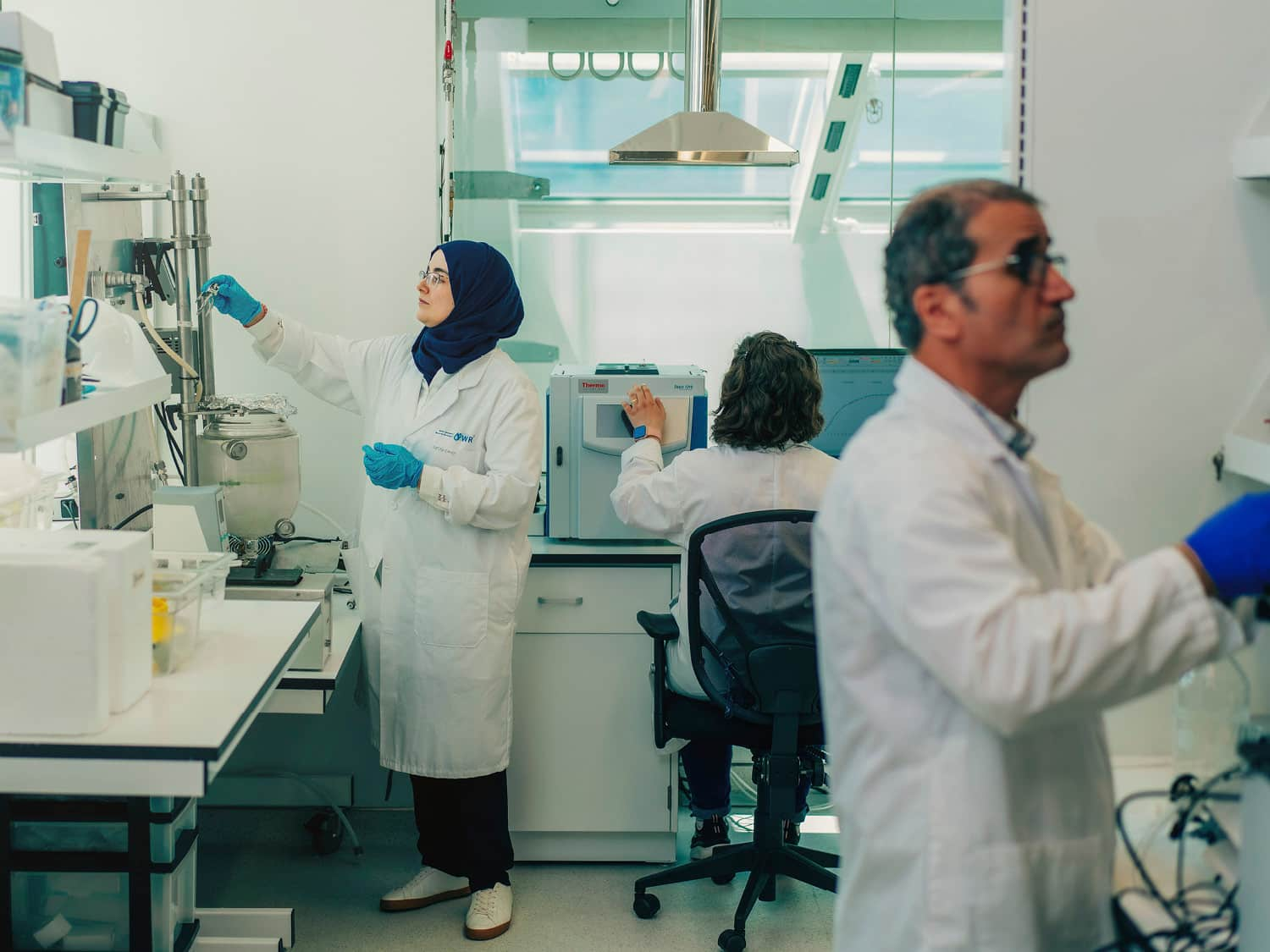 Three scientists work in a clean, white lab. A woman in a hijab and lab coat inspects a piece of equipment, while a man in safety goggles works in the foreground, and another person works at a computer in the background.