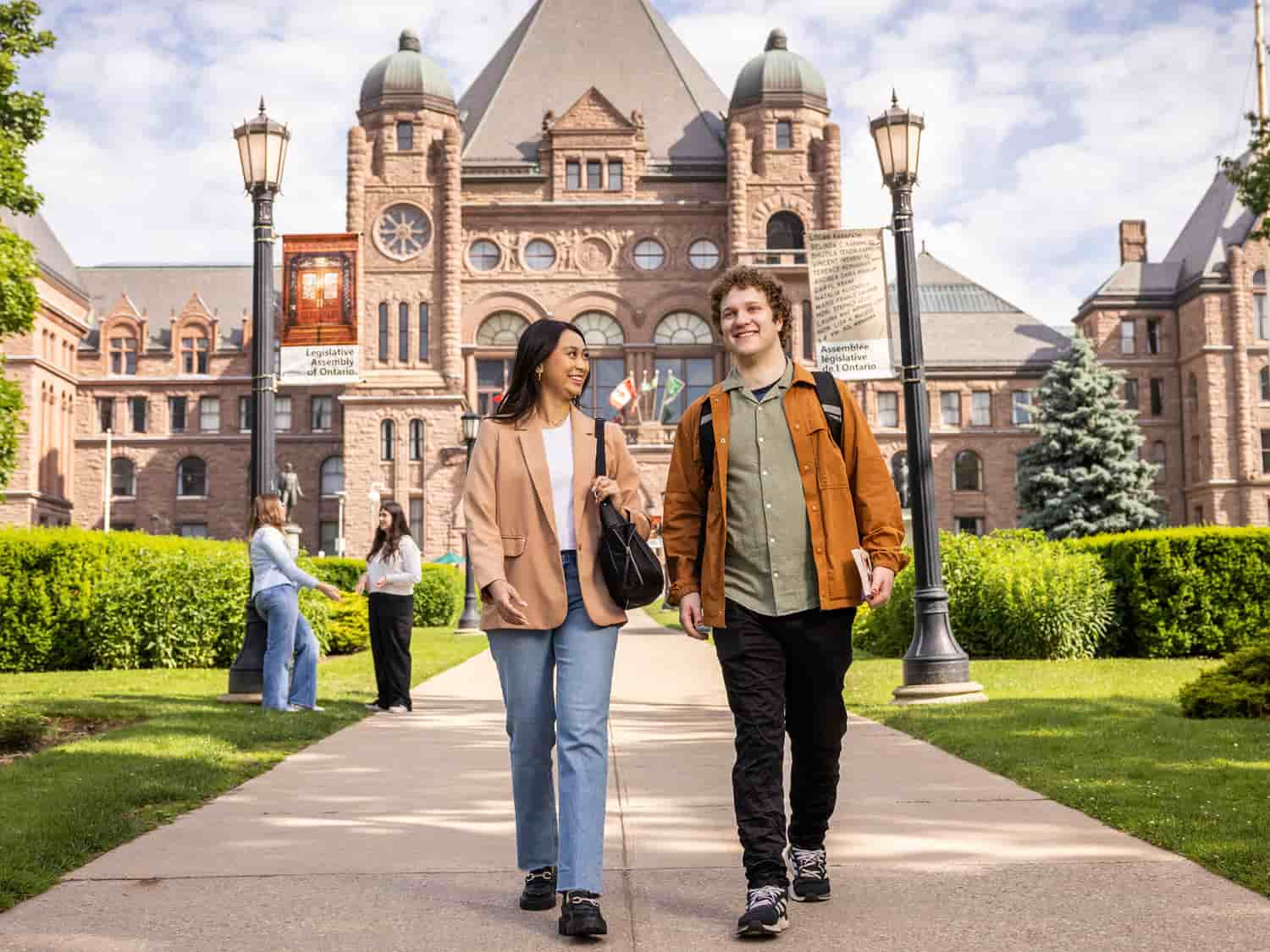 Two smiling students walk together on a path outside a historic building with stone architecture, likely on a university campus. Other students interact in the background on a sunny day.