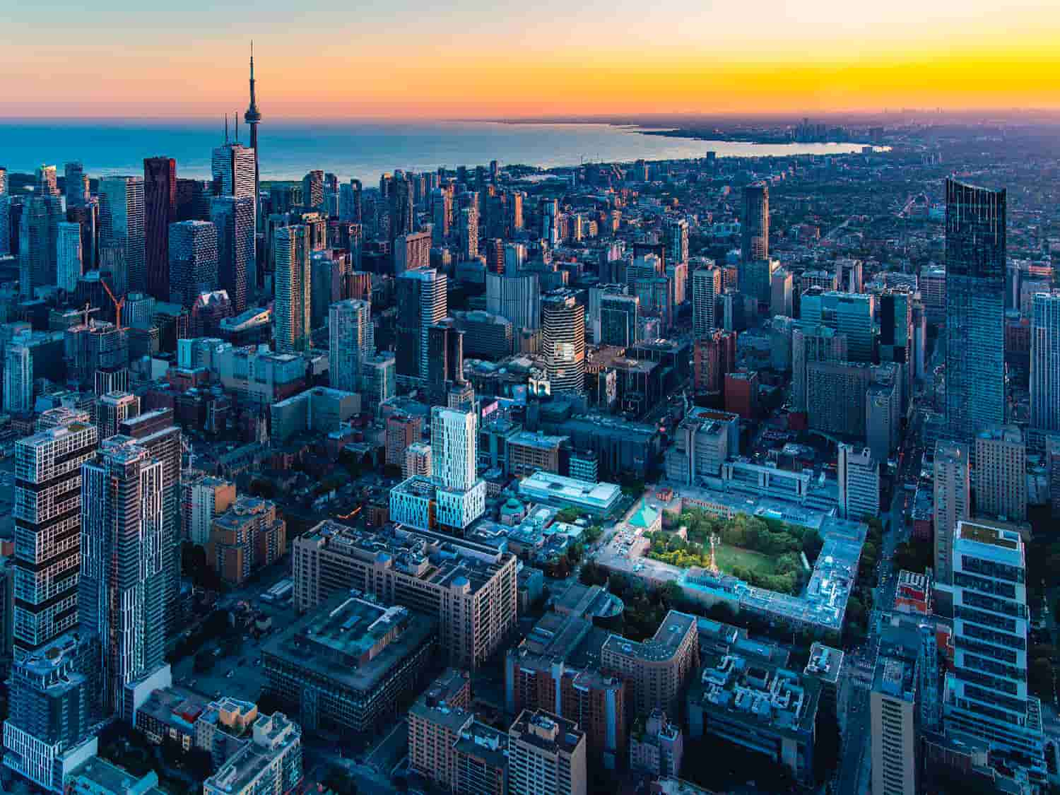 Aerial view of a cityscape at sunset, featuring skyscrapers, urban buildings, and a prominent tower, with a body of water in the distance.