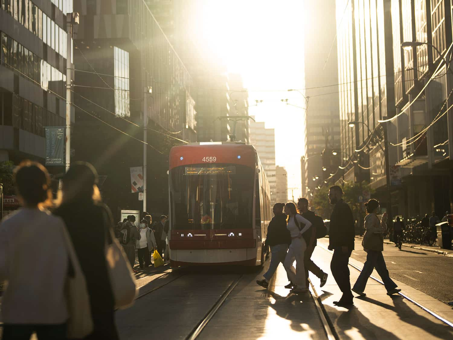 A city street scene with pedestrians crossing in front of a red streetcar during sunset, casting long shadows on the road as warm light filters through tall buildings.