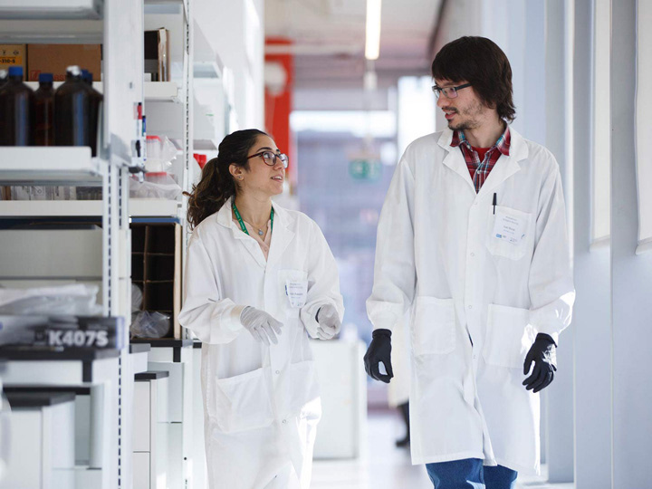 Two scientists in lab coats walking and talking in a well-lit laboratory. Shelves filled with lab supplies are visible in the background, and both appear focused and engaged.