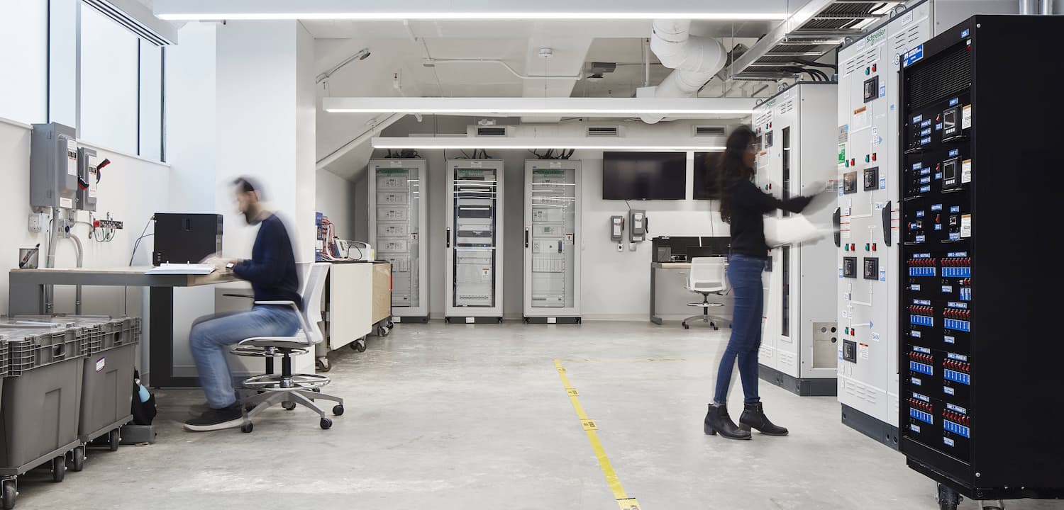 A lab with technical equipment; a seated person works at a desk while another adjusts a control panel