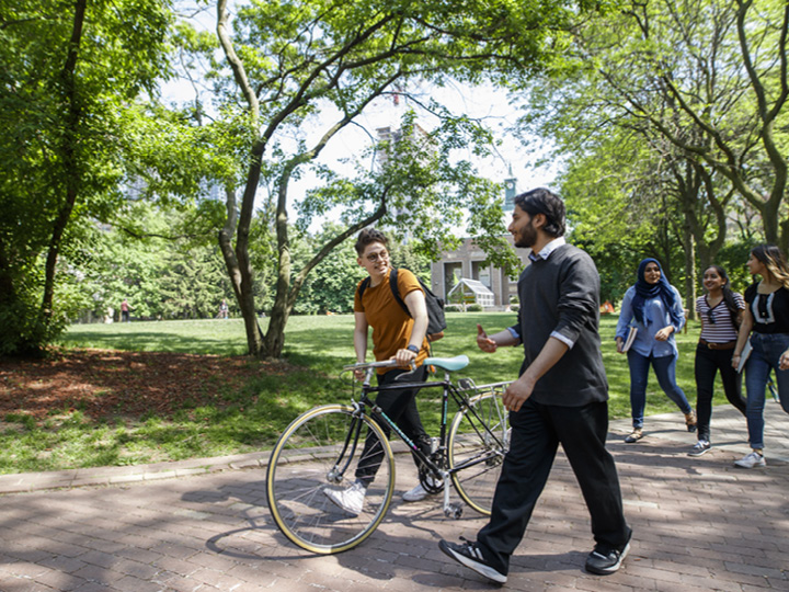 Group of students walking through a campus pathway surrounded by trees, one student pushing a bicycle, engaged in conversation.