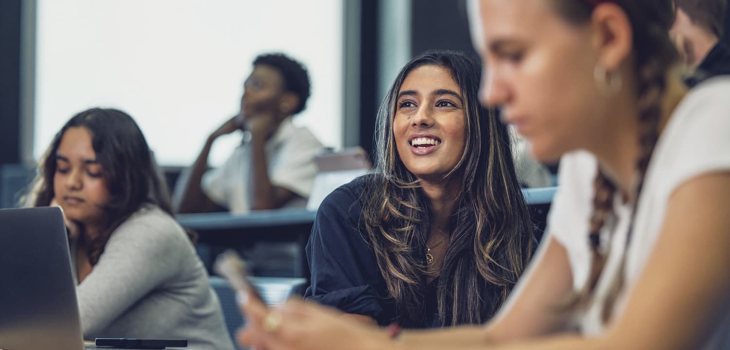 A diverse group of students is seated in a classroom setting. One student in the center of the image is smiling and appears engaged, while other students around her are focused on their own tasks, using laptops or mobile devices.