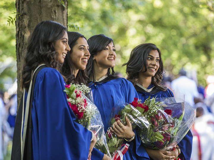 Four women in blue graduation gowns holding bouquets, smiling and standing outdoors.