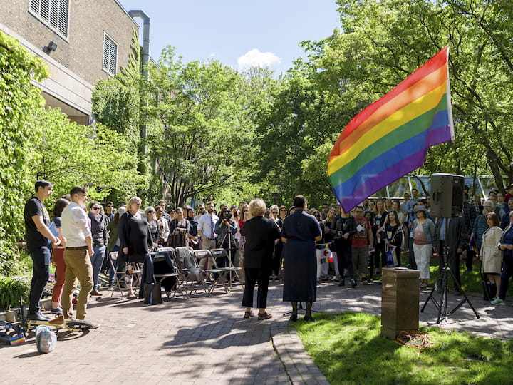 A pride flag waves vibrantly in the foreground. In the background, a group of people stand closely together, facing away from the camera.