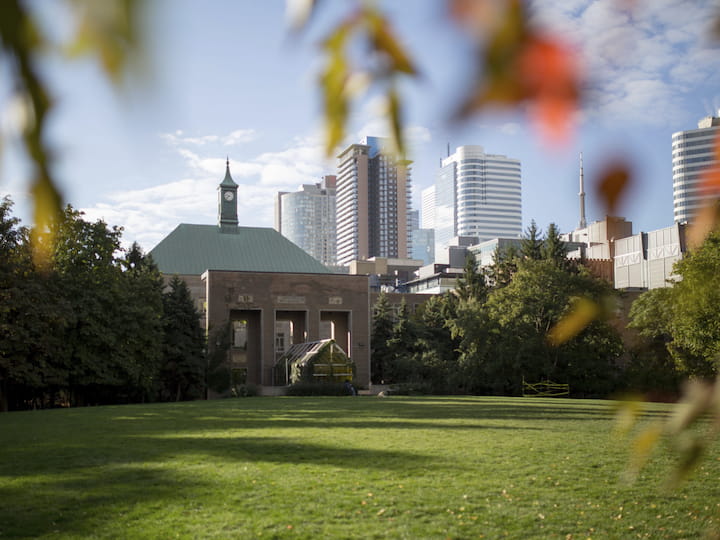 A clock tower building stands in the center of a green, open lawn, surrounded by trees and framed by leaves in the foreground. Tall modern skyscrapers rise in the background under a blue sky.