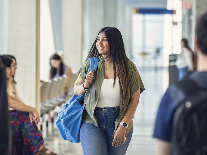 A student walks confidently down a bright hallway, carrying a large blue bag and smiling.