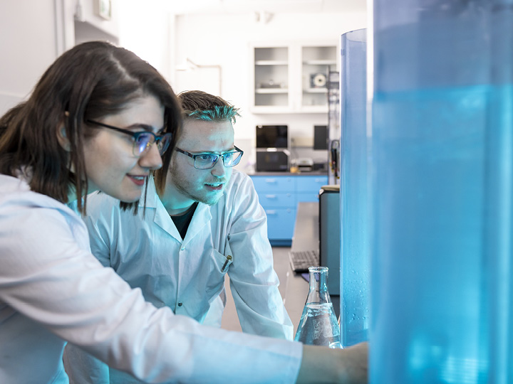 Two scientists in lab coats examining a large water tank with a beaker nearby in a lab setting.