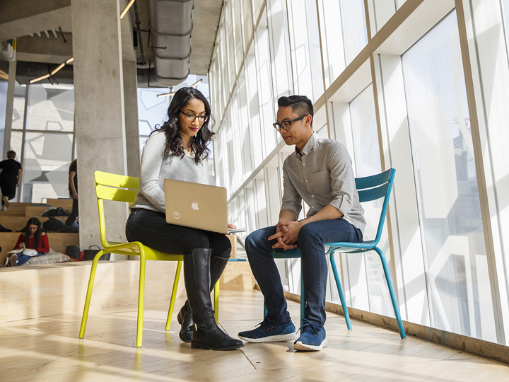 Two students sit side by side while one shows the other something on a laptop.