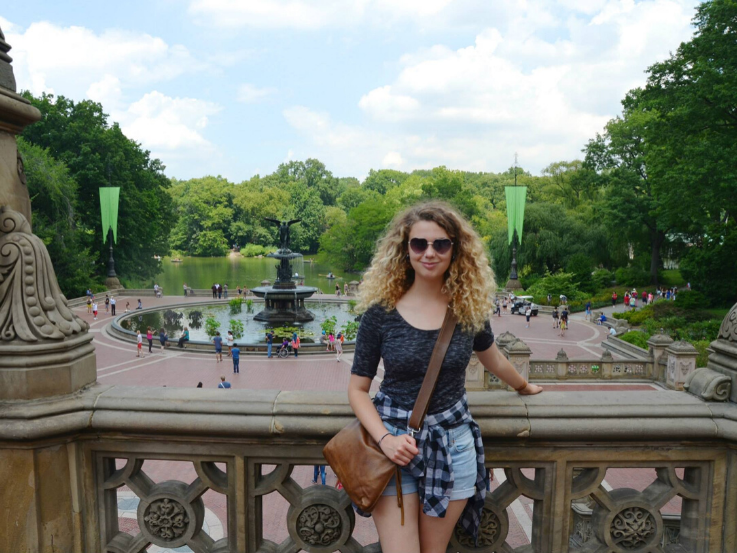 Jocelyn Courneya, wearing sunglasses, grey t shirt,  and jean shorts, standing in front of fountain in Central Park. 