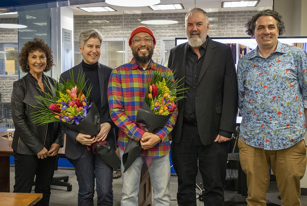 A group of 4 people standing in front of the Ryerson University Archives. Two of the people are holding colourful bouquets of flowers.