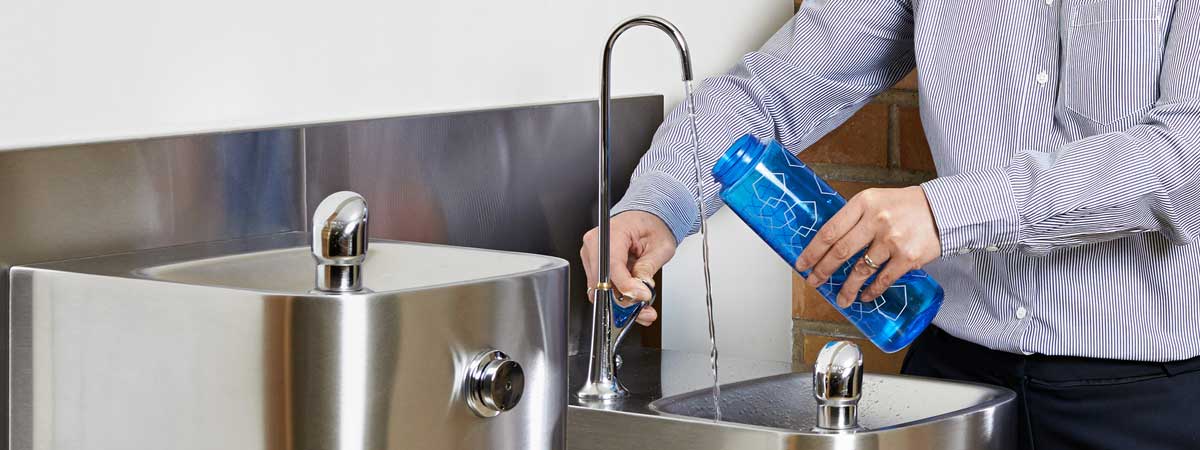 A person holding a refillable blue water bottle to the spout of a water fountain. 