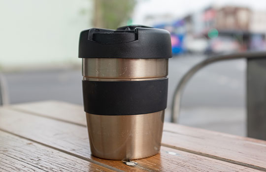 A stainless steel reusable coffee mug with a black grip and black top.