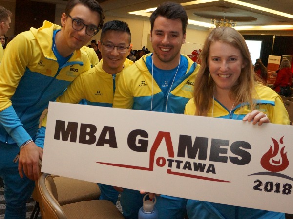 A team of four students in yellow and team zip up hoodies holding a sign at the MBA games in ottawa