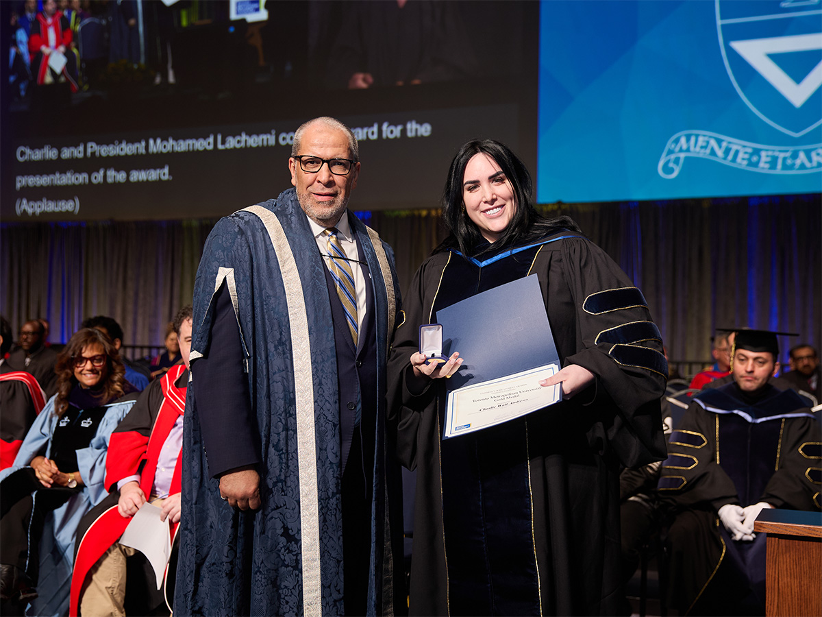 Charlie Wall-Andrews stands on stage wearing a graduation gown next to President Mohamed Lachemi as she receives Gold Medal Award