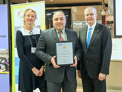 Three faculty members posing with a framed certificate in front of an open concept kitchen at an awards ceremony