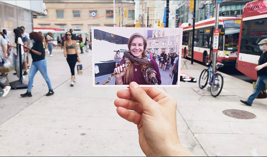 A hand holds up a printed photo of a girl with a large smile and a snake loosely wrapped around her neck. The backdrop behind the printed photo is a busy downtown street.