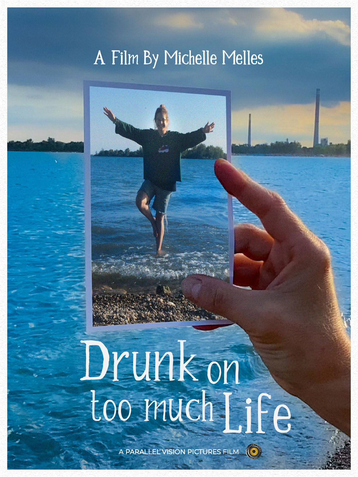 A hand holds a printed photo of a girl jumping on a lake shore. The printed photo is held against the backdrop of a similar looking lake shore.