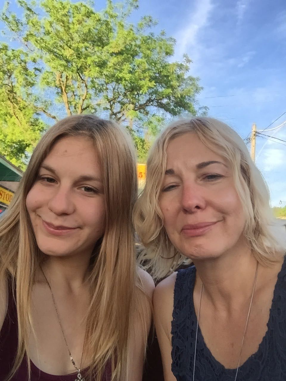 A mother and daughter taking a selfie together against the backdrop of a sun kissed tree and vivid blue sky.