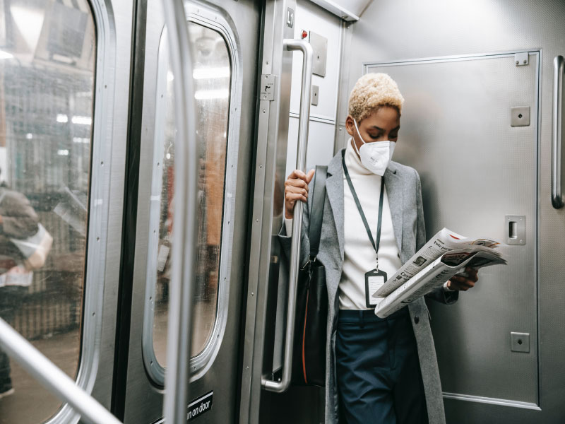 Young woman wearing a face mask reading a newspaper as she holds onto a handle inside a subway car