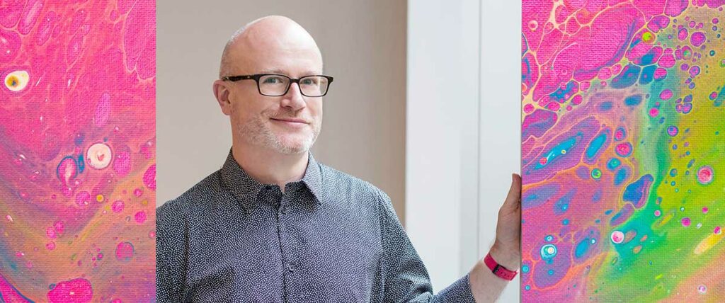 Dr. David Gauntlett standing with his hand on a wall, smiling. He wears eyeglasses and collared dress shirta 