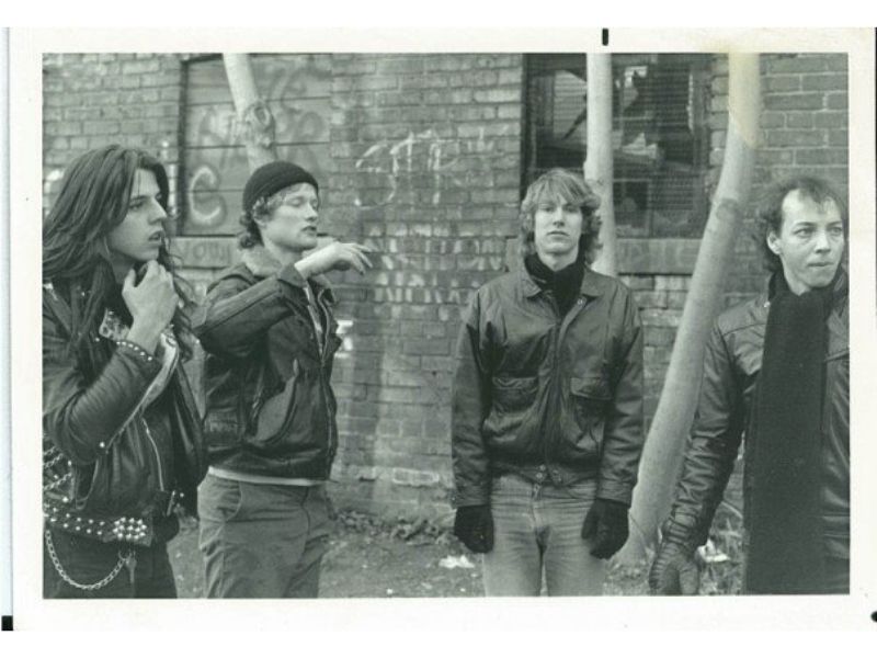 Four young men wearing black leather jackets in front of a brick wall covered in graffiti.