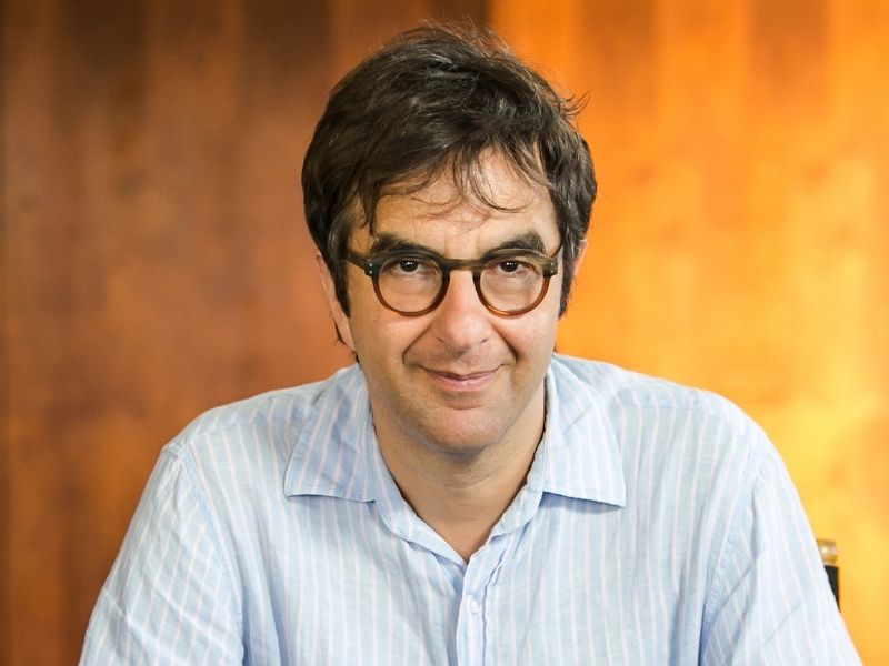  Headshot of Egoyan. He has dark hair and wears dark-cloured glasses with a collared shirt. He sits in front of a graded orange and yellow background