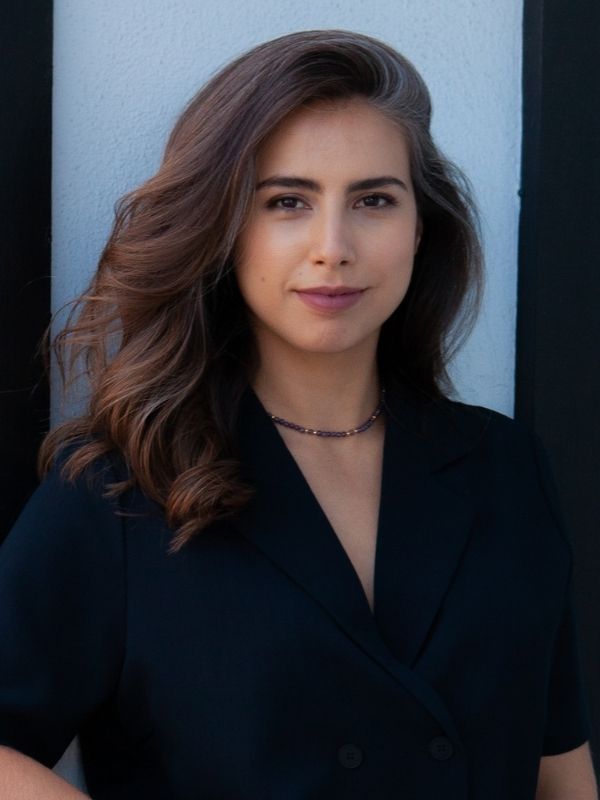 Portrait of Nansy Khanano. She wears a black v-neck shirt and a beaded necklace. She smiles with her mouth closed to the camera. Khanano has brown hair and leans against a light blue wall.