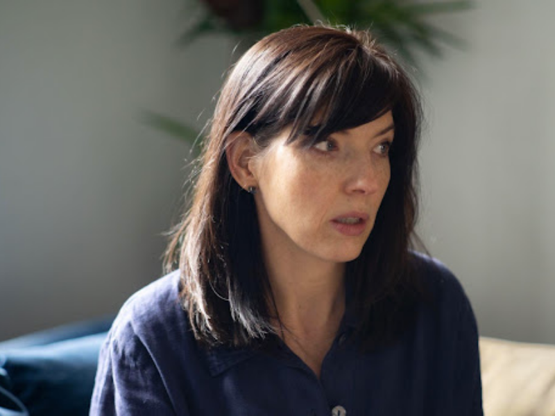 A woman staring off to the right side of view. Her long, dark hair is draped over a blue button-up and a plant and white wall is blurred in the background.