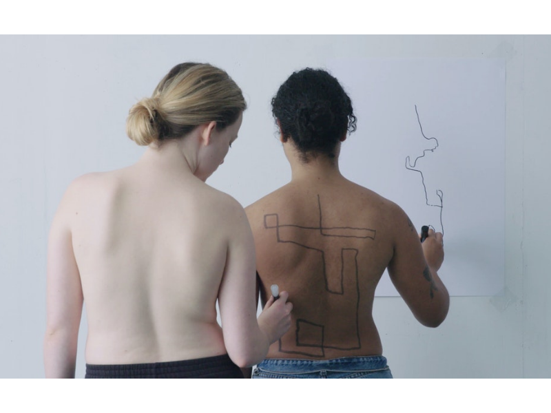 Two women facing away from the camera. One is drawing an abstract line drawing with a sharpie on a white wall while the other is drawing a similar shape on the others back.